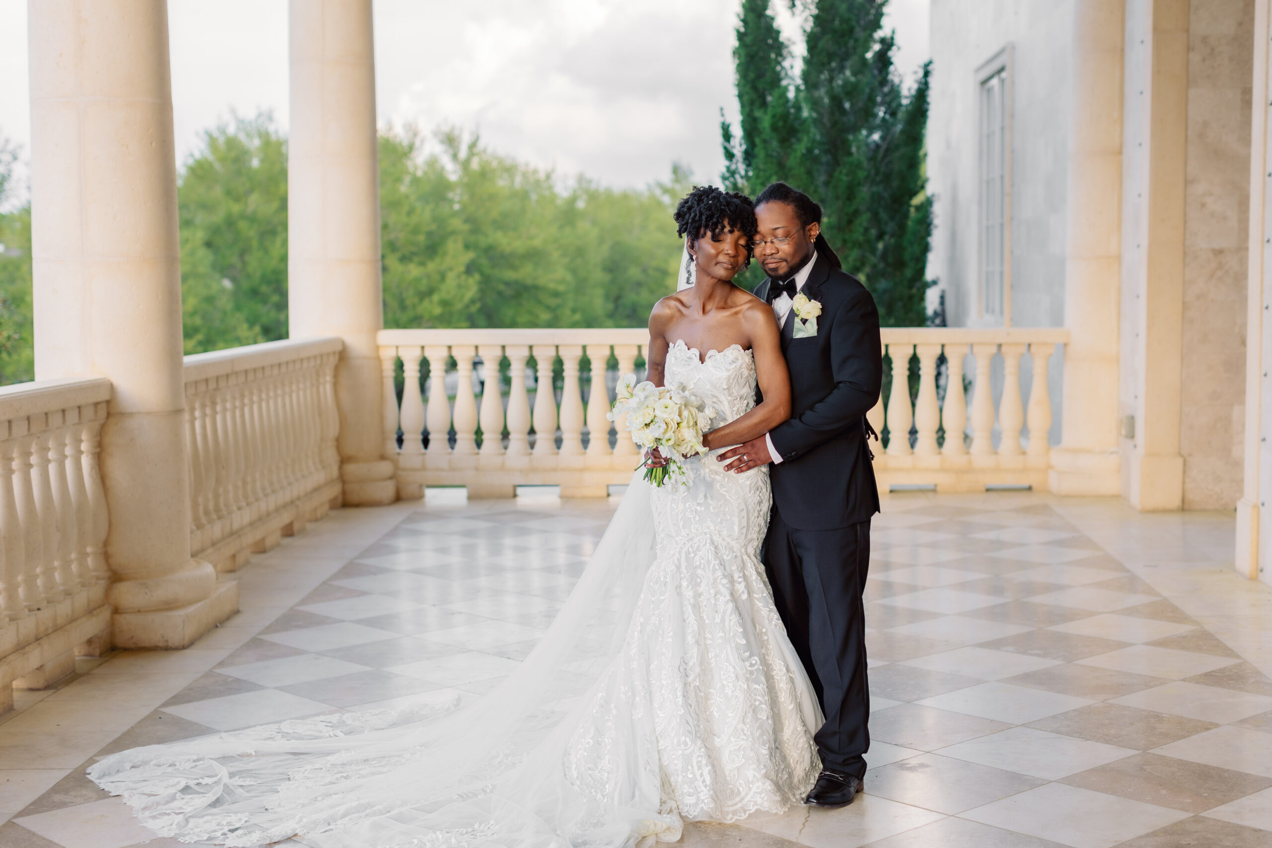 Michelle and Deondre, the newlywed couple, embracing on the veranda of their wedding venue at The Regent. Michelle wears a white gown with intricate details, a long train, and a veil, holding her bouquet. Deondre is in a black tux.